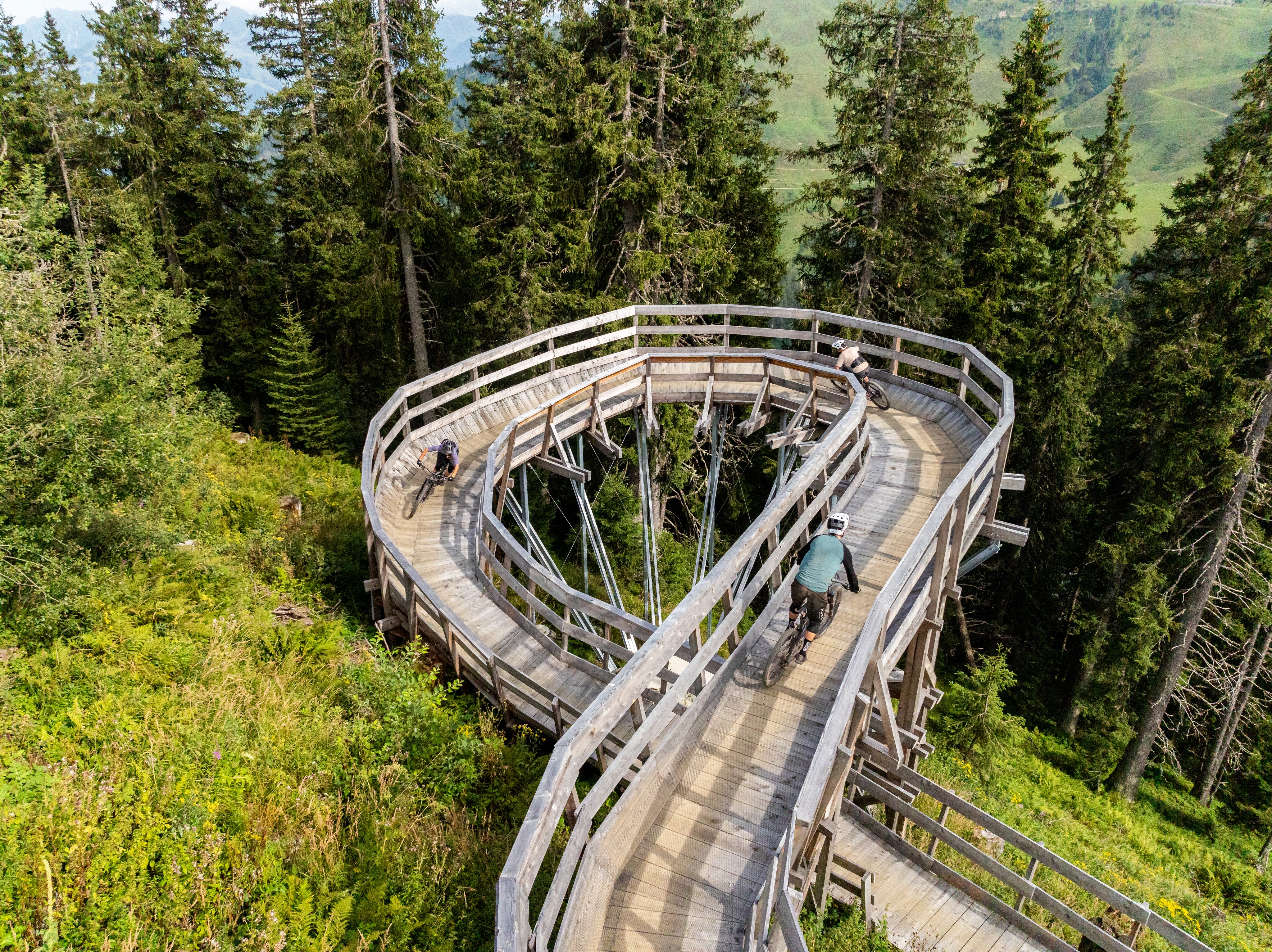 Die Aufnahme zeigt eine hölzerne Kurvenkonstruktion für Mountainbiker im Wald. Mehrere Fahrer nutzen die Rampe als Teil einer Downhill-Strecke.