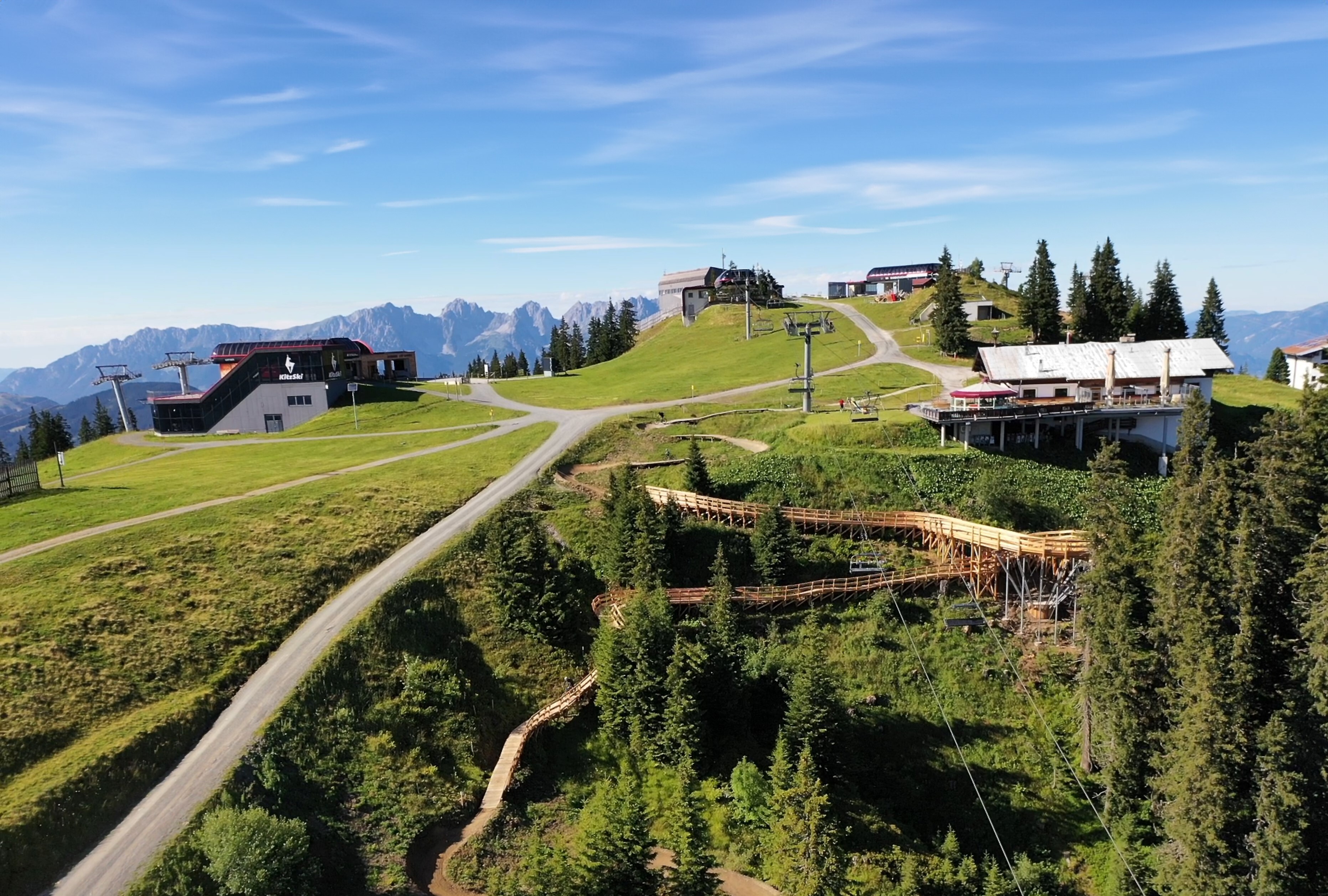 Die Aufnahme zeigt die Bergstation der Fleckalmbahn und den Sonnenrast Trail mit seiner markanten Holzkonstruktion. Im Hintergrund sind Bergketten unter blauem Himmel sichtbar.