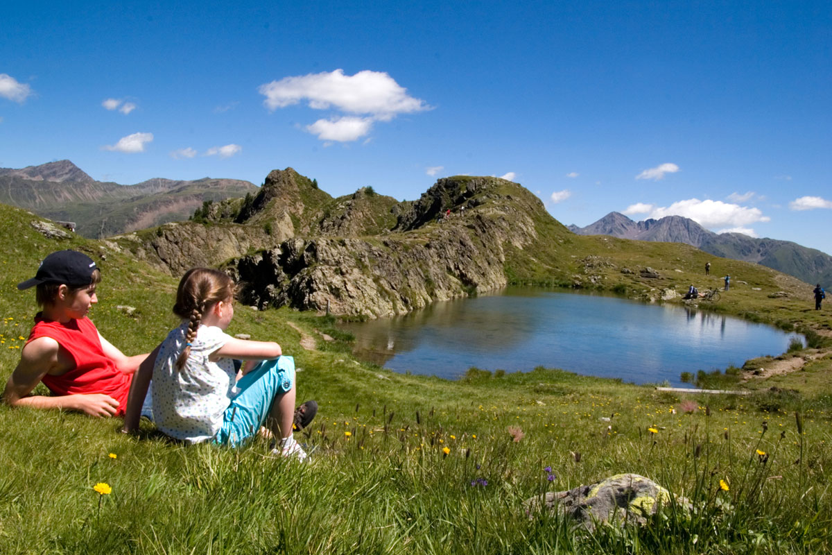Zwei Kinder rasten auf einer Bergwiese am Ufer eines der drei Seen am Thurntaler. Im Hintergrund sind felsige Berggipfel unter blauem Himmel zu sehen.