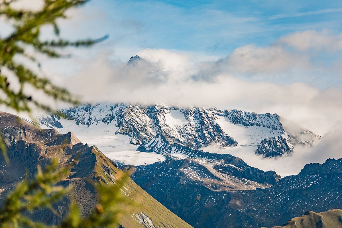 Fernsicht auf die vergletscherten Gipfel der Hohen Tauern. Das Bild zeigt alpine Felsformationen und Schneefelder unter einer Wolkendecke am Adler Trail.