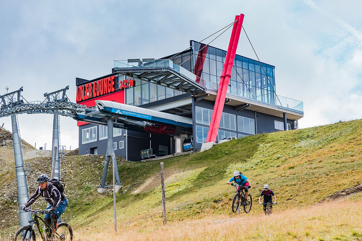 Mountainbiker befahren den Adler Trail Nr. 138 im alpinen Gelände. Im Hintergrund ist die moderne Architektur der Adler Lounge mit der angrenzenden Seilbahnstation zu sehen.