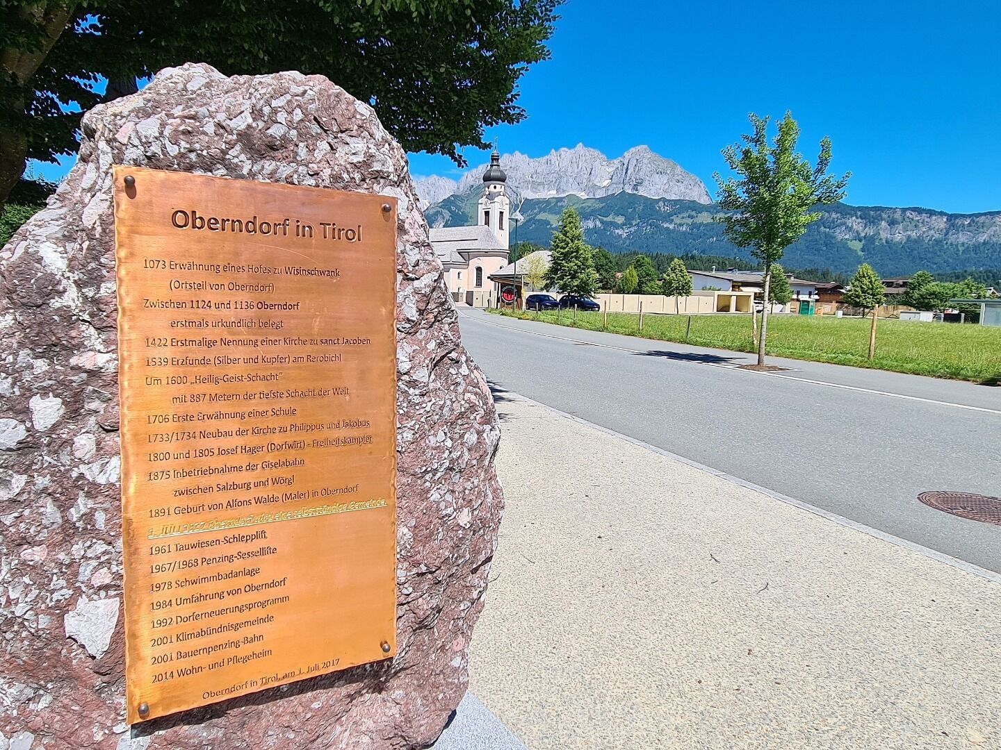 Eine Informationstafel zur Ortsgeschichte von Oberndorf in Tirol ist auf einem Stein montiert. Im Hintergrund sind die Dorfkirche und das Bergmassiv des Wilden Kaisers zu sehen.
