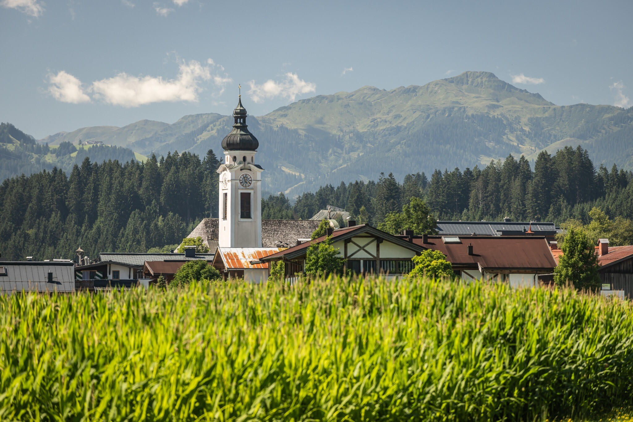Die Aufnahme zeigt die Ortschaft Oberndorf in Tirol mit ihrer markanten Kirche. Im Hintergrund erstreckt sich die bewaldete Berglandschaft unter leicht bewölktem Himmel.