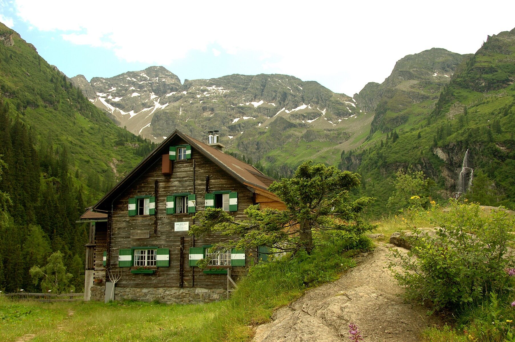 Die Hans-Wödl-Hütte ist eine traditionelle Schutzhütte aus Holz. Sie liegt eingebettet in eine Berglandschaft mit steilen Hängen und einem sichtbaren Wasserfall im Hintergrund.