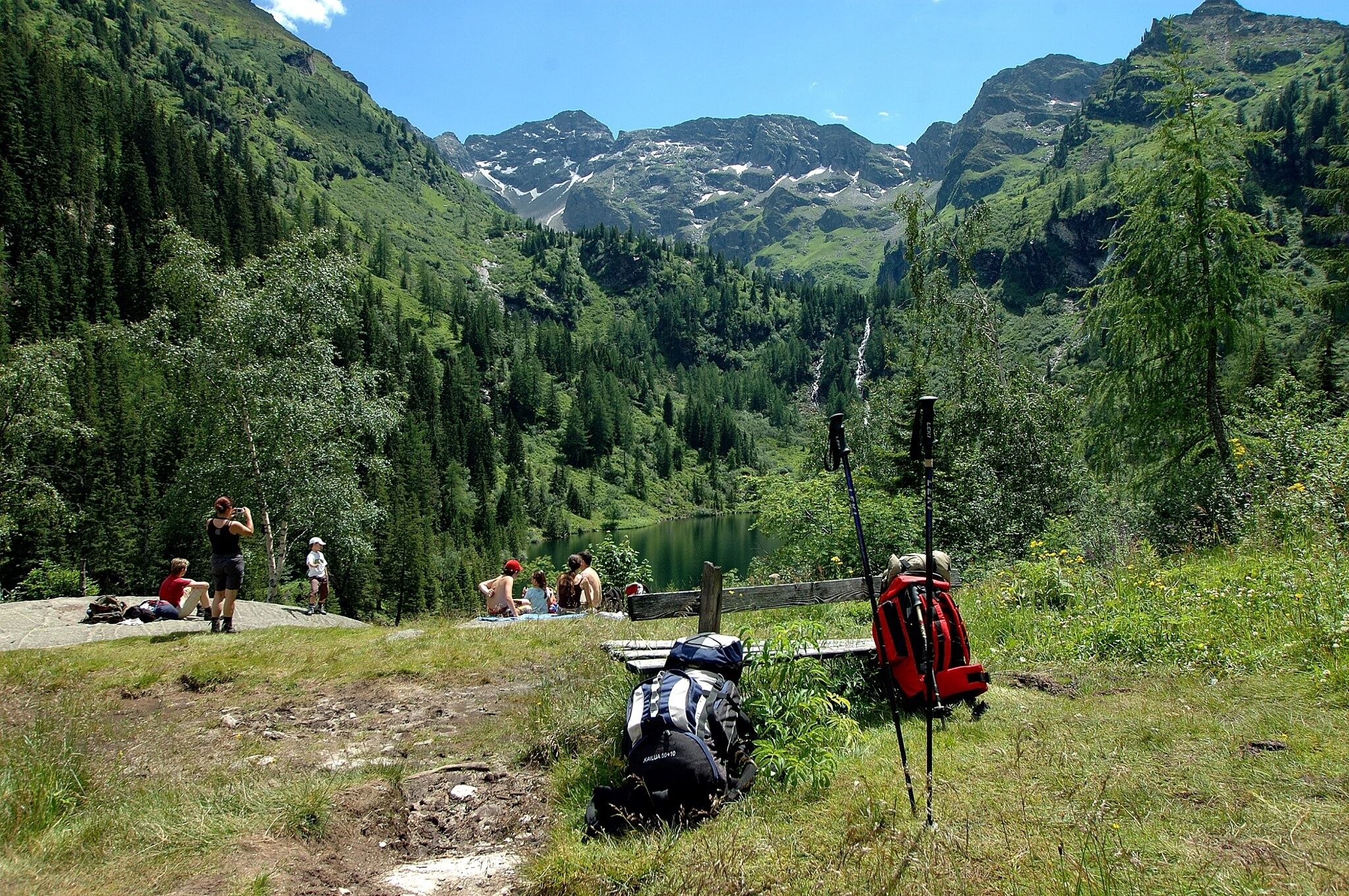 Wanderer machen Pause auf einer Bergwiese. Im Hintergrund sind der Hüttensee, dichte Nadelwälder und felsige Berggipfel zu sehen.