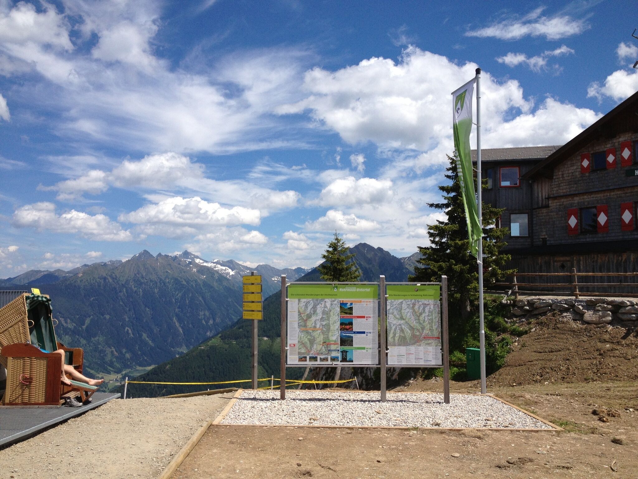 Eine Informationstafel mit Wanderkarten steht neben einer Berghütte. Im Hintergrund sind die Gipfel der Alpen unter leicht bewölktem Himmel zu sehen.