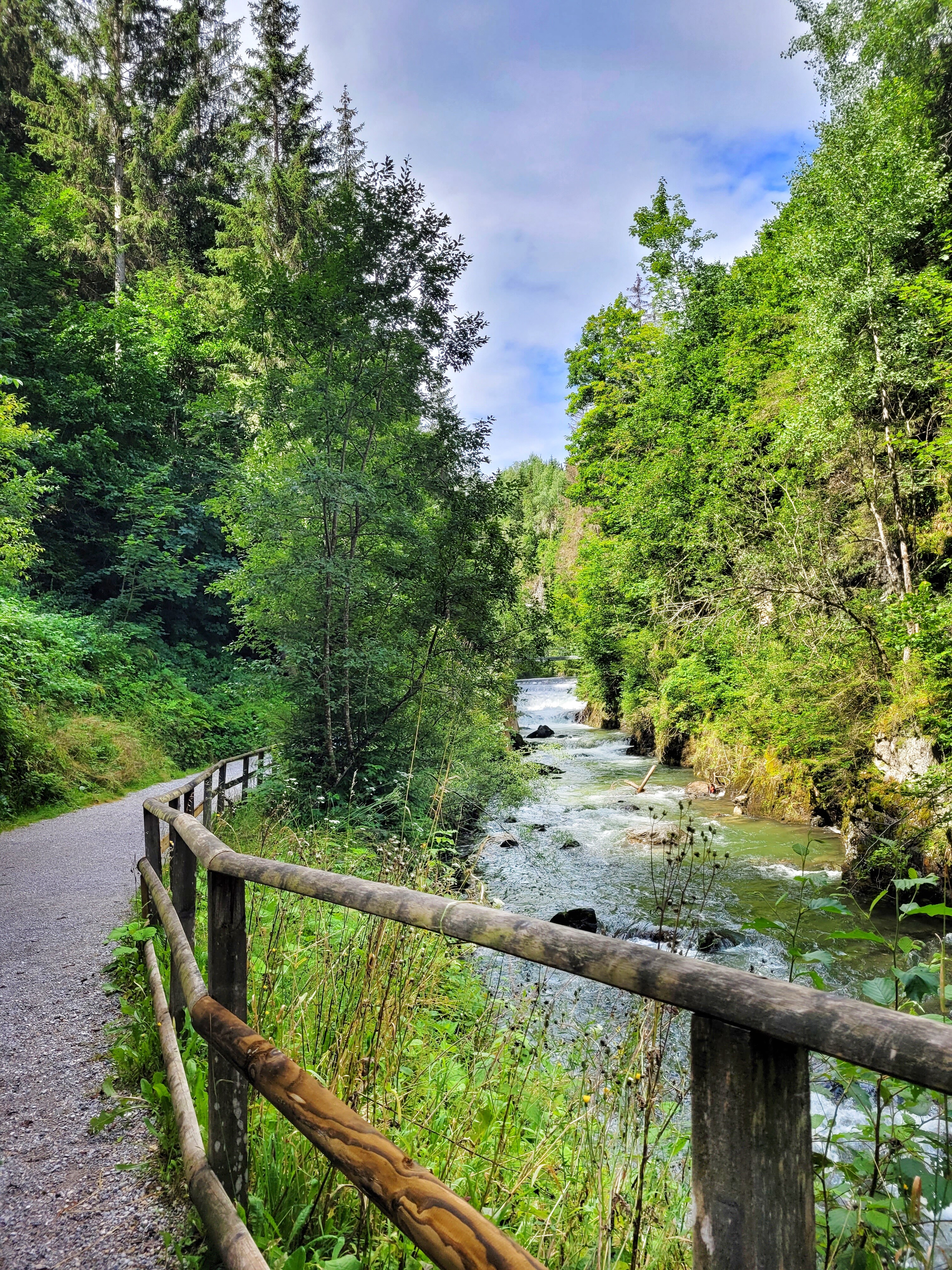 Der befestigte Wanderweg führt entlang des Talbachs durch bewaldetes Gebiet. Ein Holzgeländer trennt den Weg vom fließenden Gewässer.