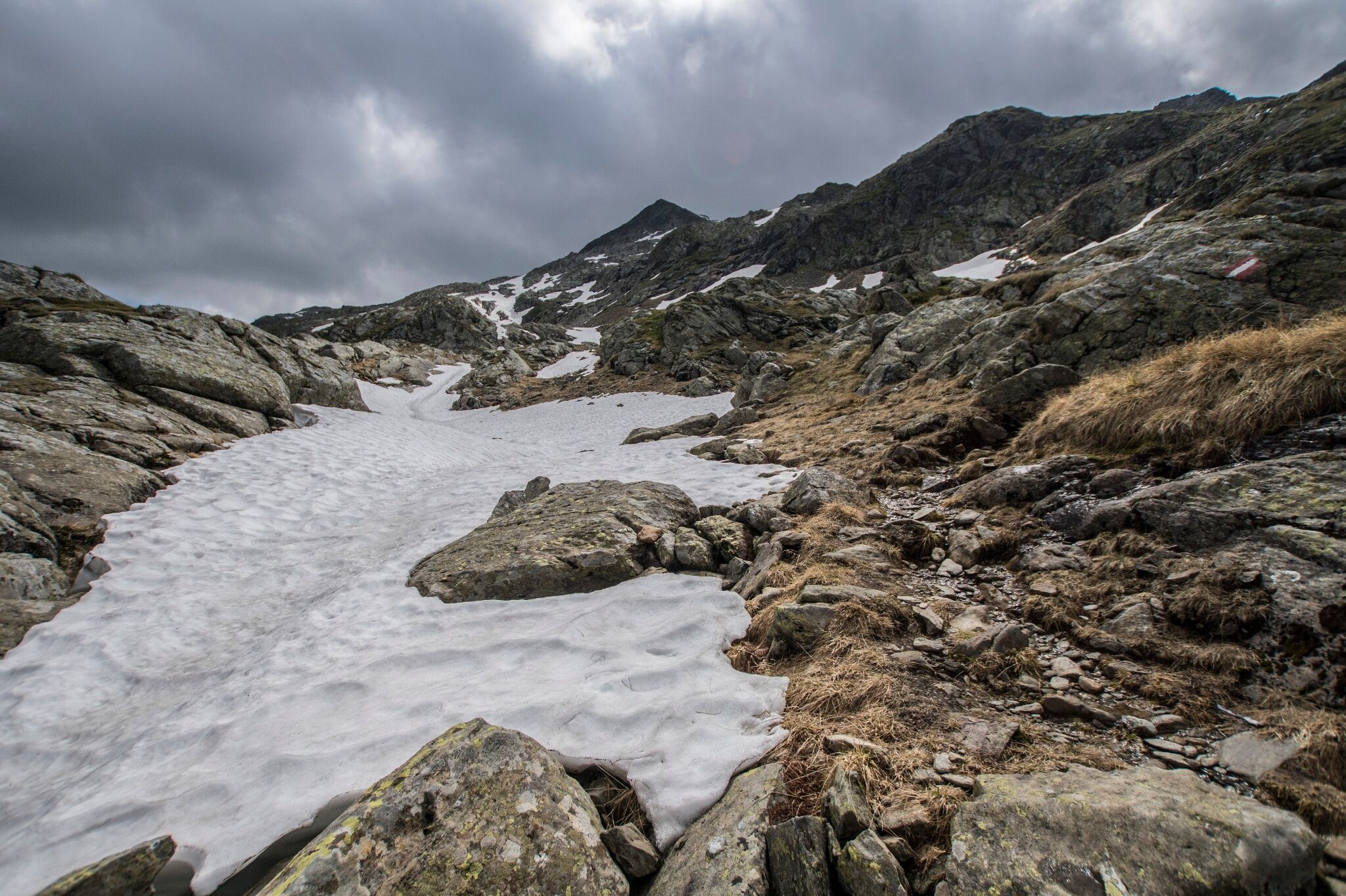 Blick auf ein Schneefeld in einer steinigen Hochgebirgslandschaft. Im Hintergrund ist die Silhouette der Vetternspitze unter Wolken zu sehen.