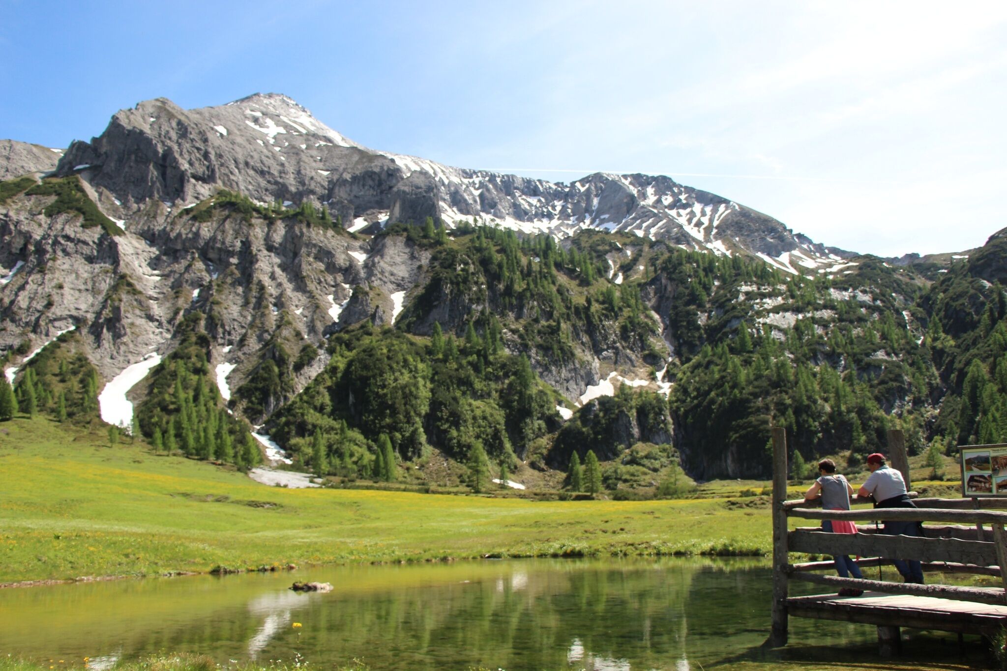 Wanderer blicken von einem Holzsteg auf einen kleinen See im Almgebiet. Im Hintergrund ragt die Steirische Kalkspitze mit ihren markanten Felswänden auf.