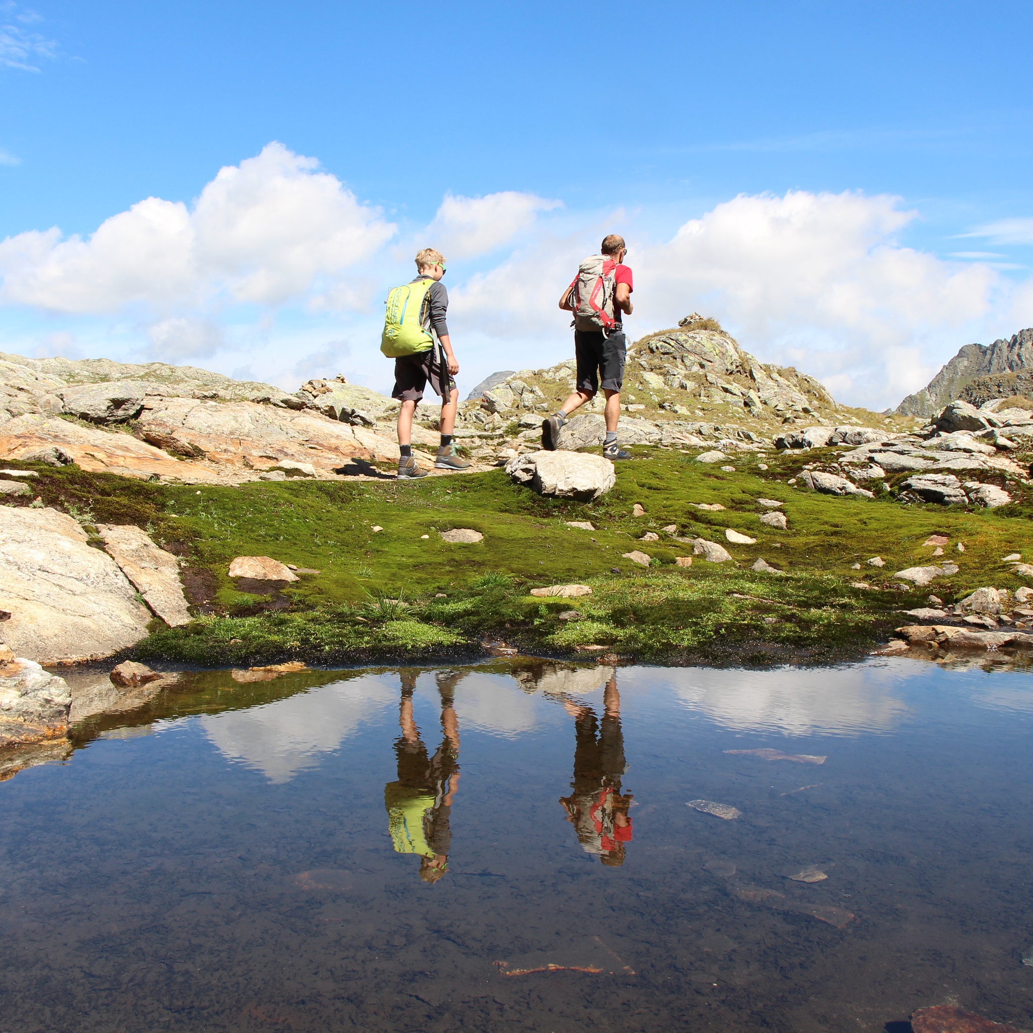Zwei Personen wandern durch alpines Gelände an einem kleinen Bergsee. Die Wasseroberfläche spiegelt die Wanderer und den bewölkten Himmel wider.