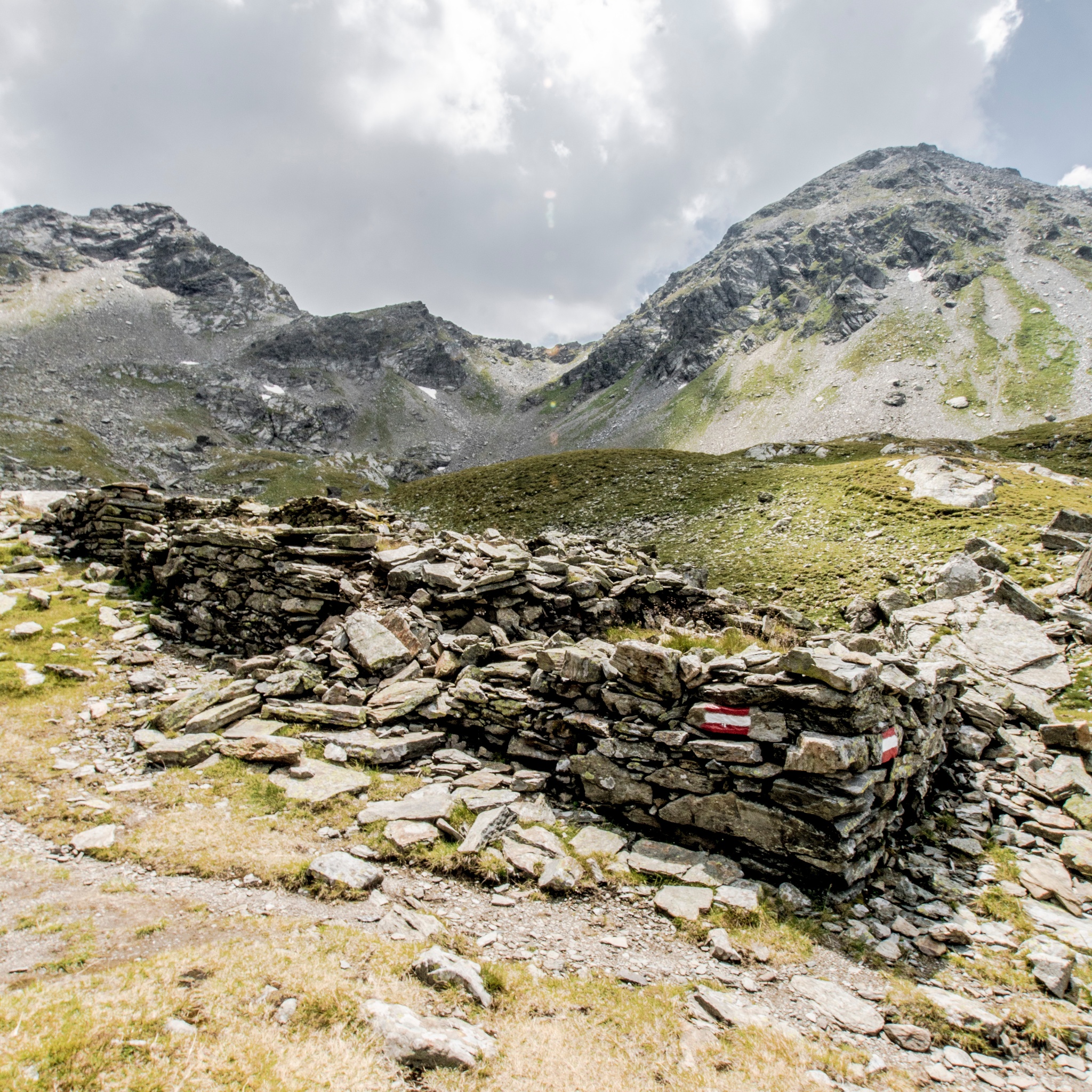 Überreste einer ehemaligen Knappenbehausung aus geschichteten Steinen im hochalpinen Gelände. Ein markierter Wanderweg führt direkt an der Ruine vorbei.