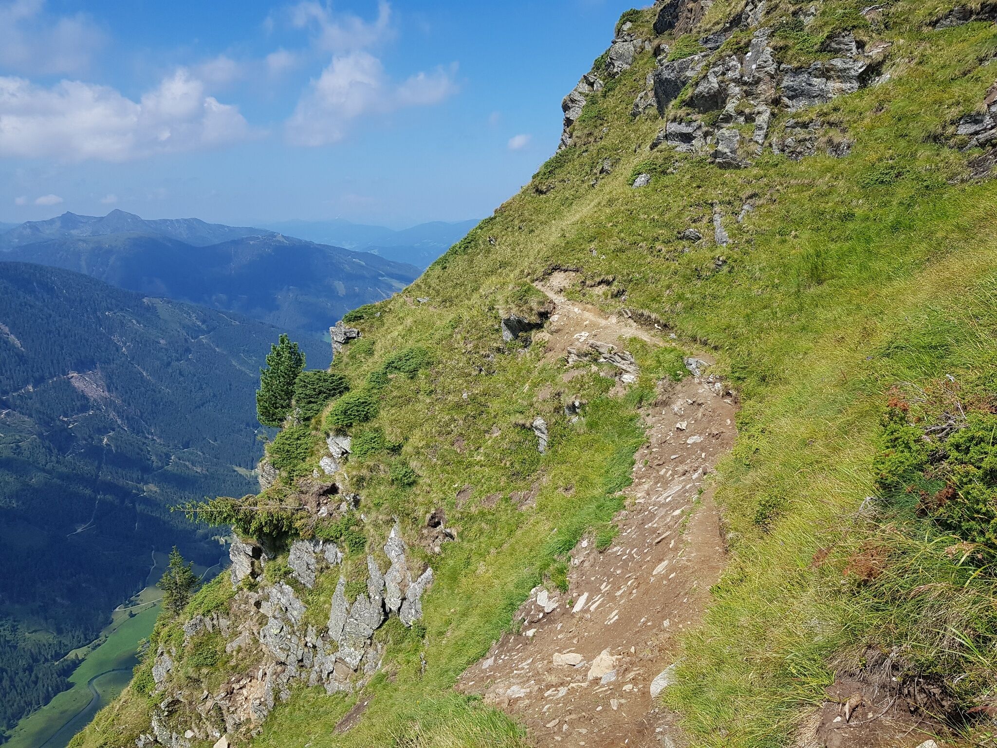 Ein schmaler Pfad verläuft entlang eines steilen Grashangs im Hochgebirge. Im Hintergrund ist ein weites Tal mit bewaldeten Bergen unter blauem Himmel sichtbar.