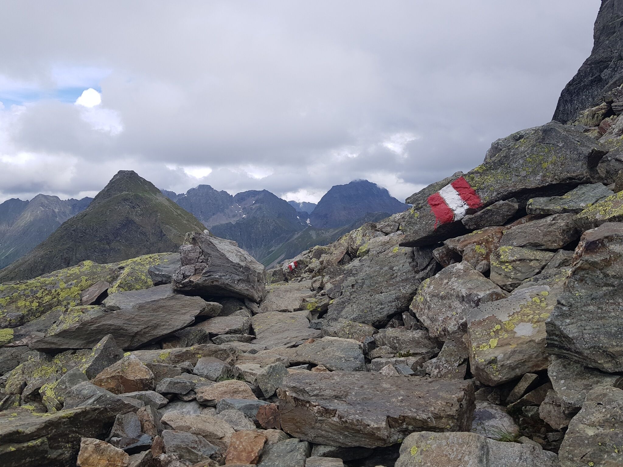 Der alpine Pfad verläuft über loses Gestein unterhalb des Sauberg. Eine rot-weiße Markierung auf einem Stein weist den Weg durch das felsige Gelände.