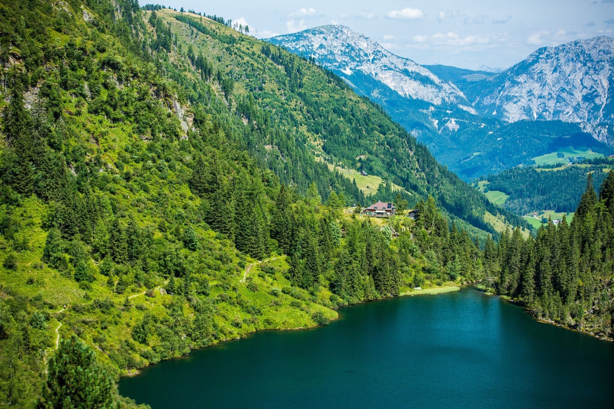 Der Hüttensee liegt eingebettet in bewaldete Berghänge. Im Hintergrund sind felsige Alpengipfel und eine bewirtschaftete Hütte zu sehen.