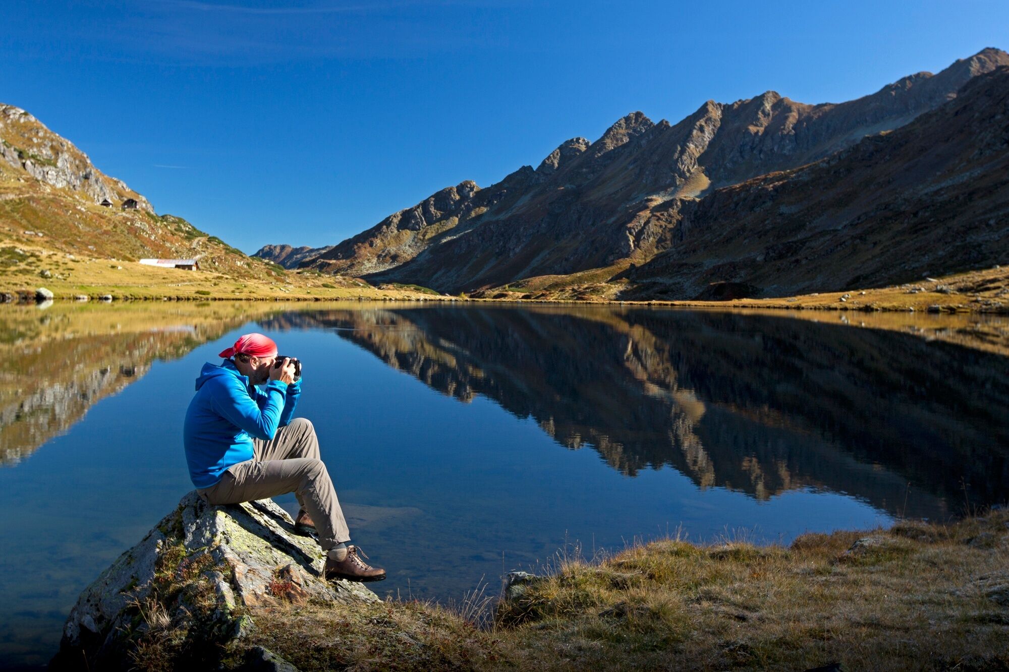 Ein Mann in blauer Jacke fotografiert die Schladminger Tauern, die sich in der glatten Oberfläche des Giglachsees spiegeln. Im Hintergrund sind felsige Berggipfel unter blauem Himmel zu sehen.
