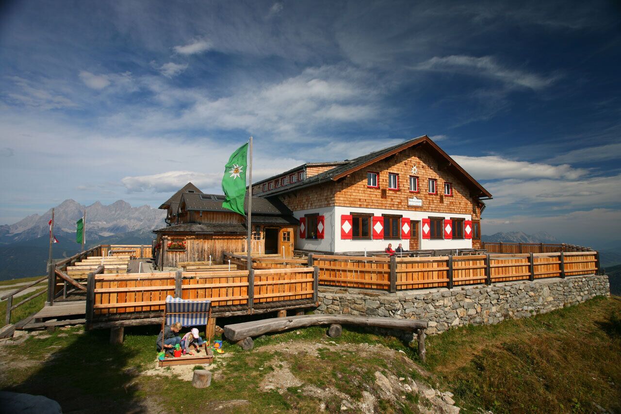 Die Hochwurzenhütte in den Schladminger Tauern verfügt über eine weitläufige Holzterrasse und einen Kinderspielbereich. Im Hintergrund sind alpine Gipfel sichtbar.