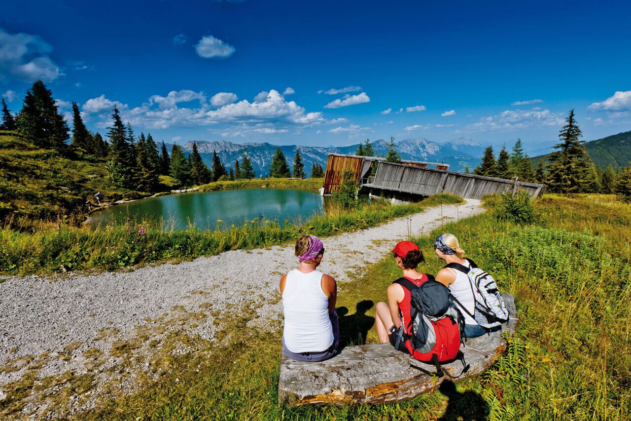 Wanderinnen rasten auf einer Bank mit Blick auf einen kleinen See und ein Holzgebäude. Im Hintergrund ist das Bergpanorama unter blauem Himmel zu sehen.