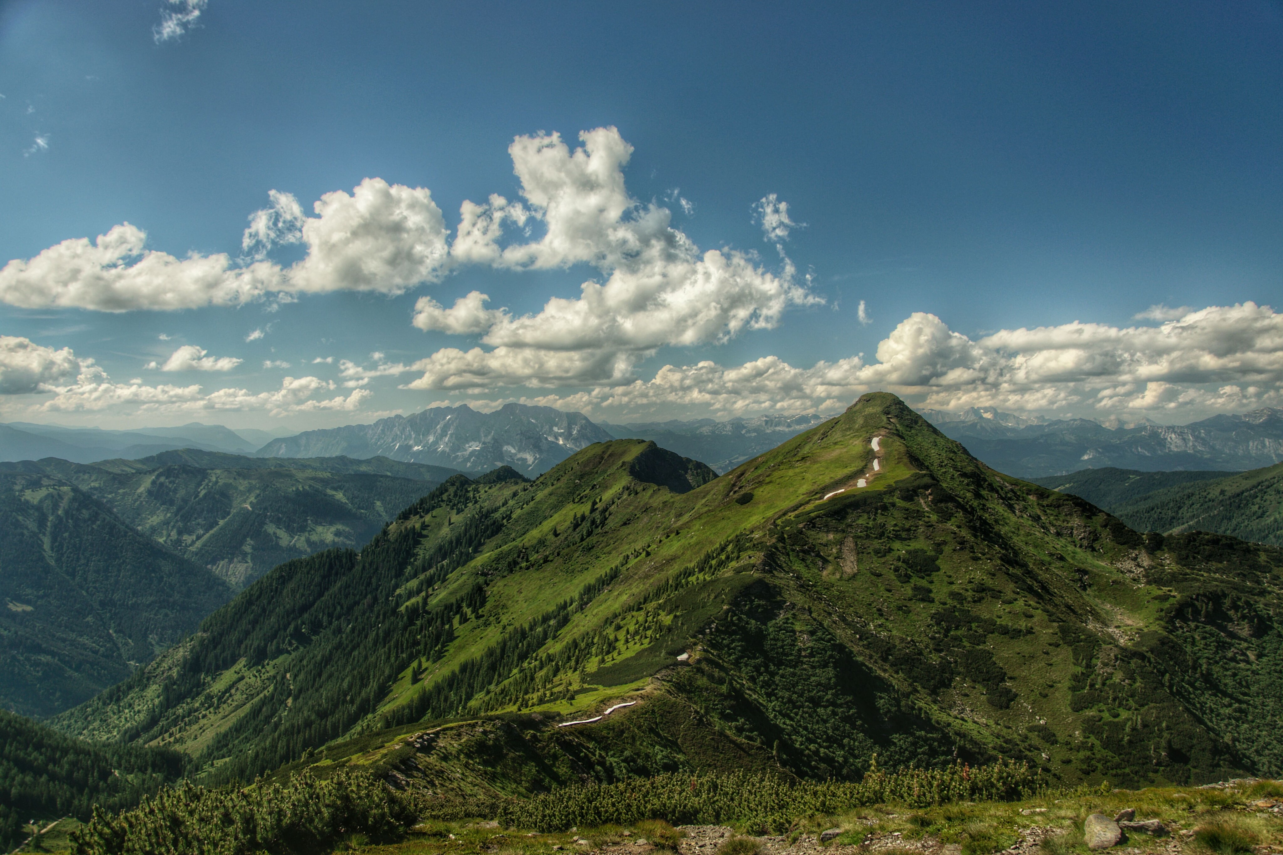 Die Aufnahme zeigt den grasbewachsenen Kamm und Gipfel des Karlspitz in den österreichischen Alpen. Im Hintergrund sind weitere Gebirgsketten und Täler unter einer lockeren Wolkendecke sichtbar.