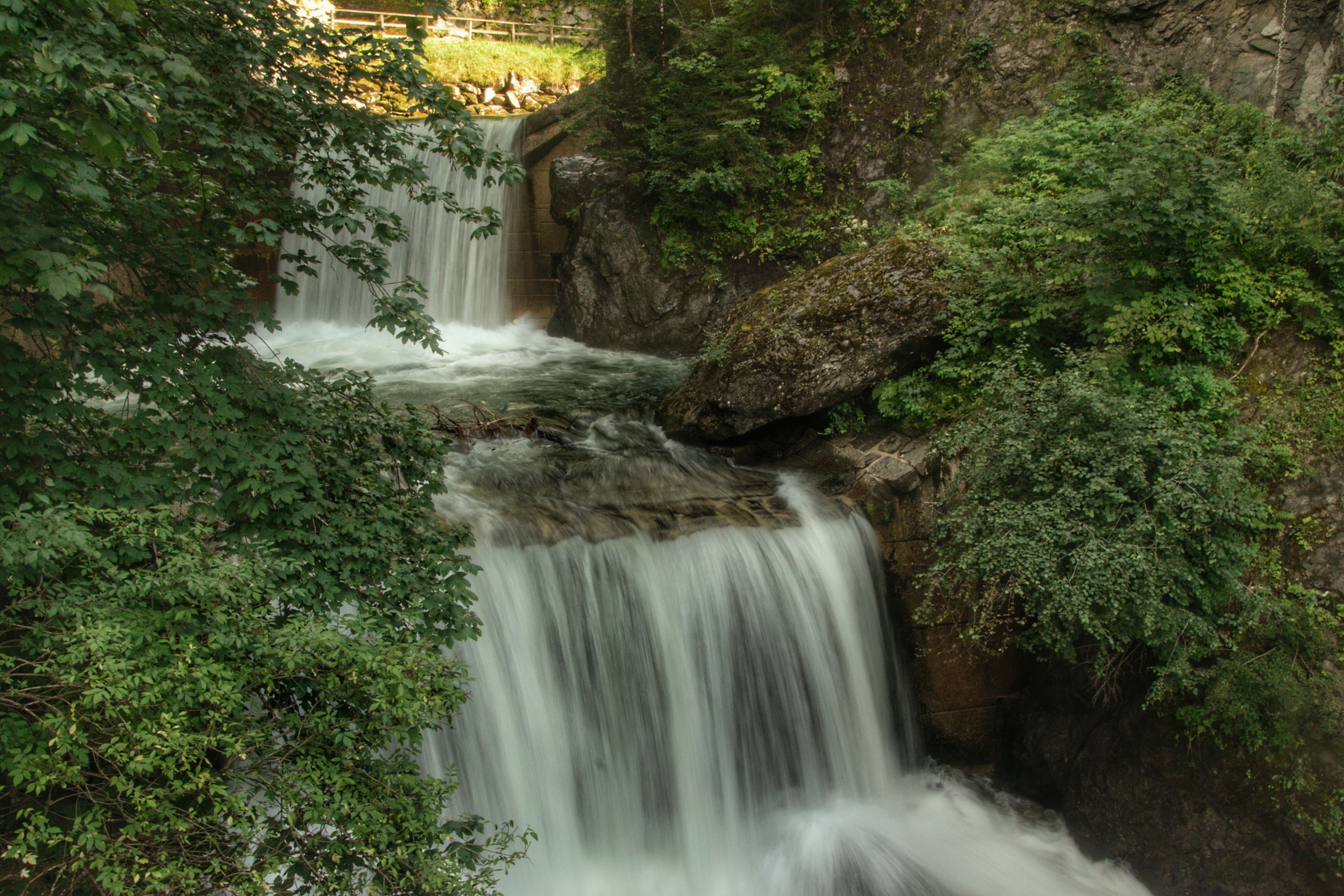 Ein Gebirgsbach stürzt über zwei Steinstufen in die Tiefe. Die Kaskaden sind von dichtem Laubwald und dunklen Felswänden umgeben.