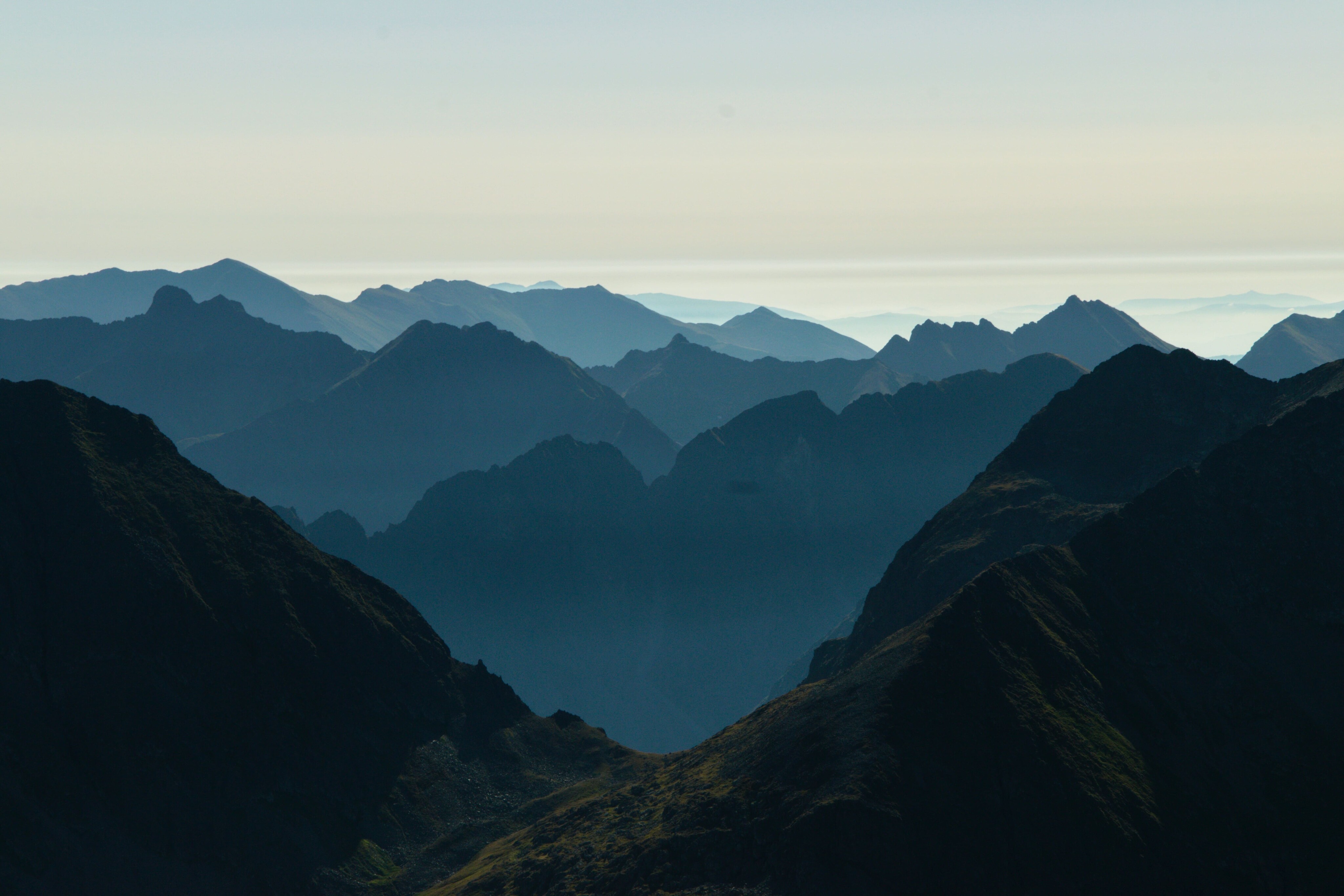 Die Aufnahme zeigt die gestaffelten Silhouetten alpiner Bergketten. Im Vordergrund sind dunkle Felsformationen zu sehen, die nach hinten in hellere Blautöne übergehen.